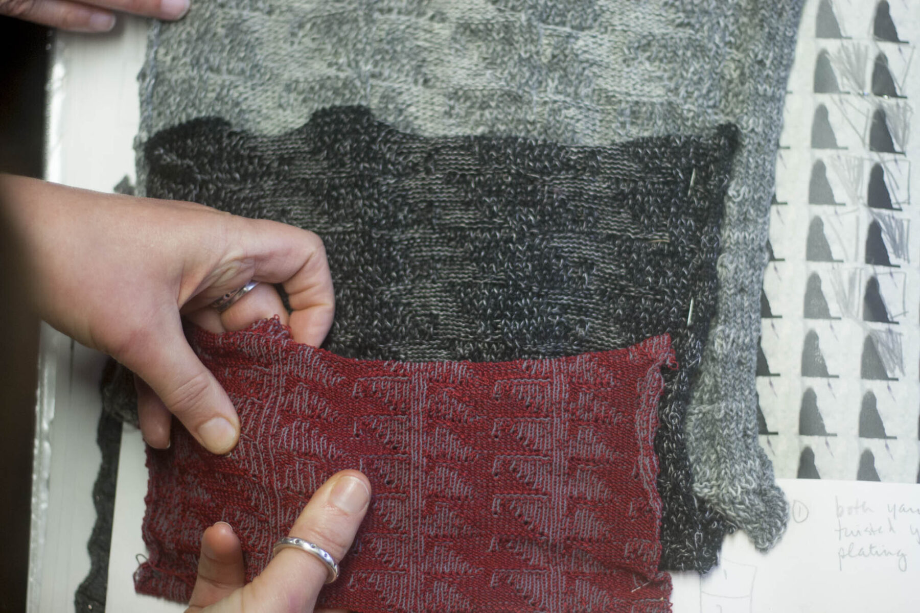 Hands exploring the stitches on Nielanell swatches. Swatches in red and grey and sitting on top of monochrome sketches.