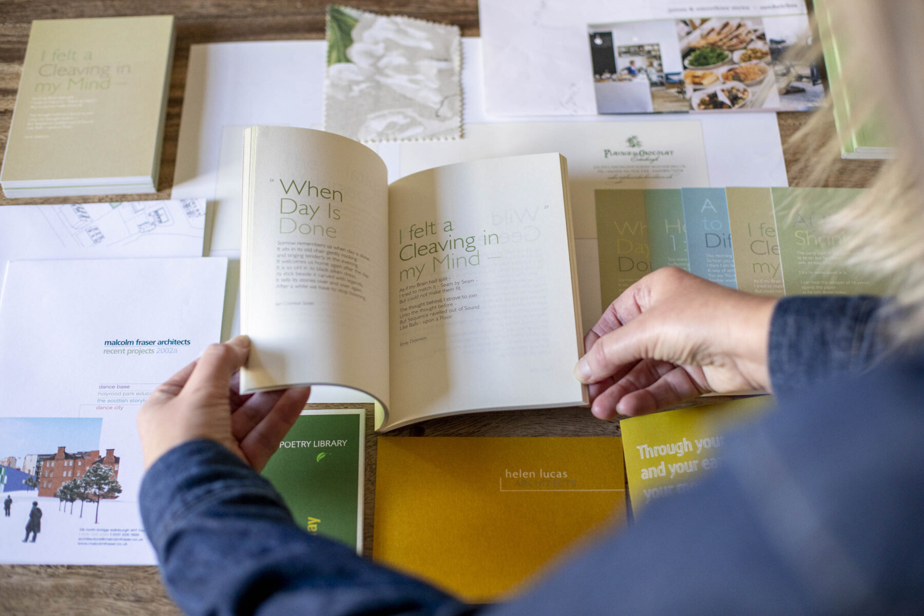 Various designed books, postcards and printed material lying on a wooden table. Hands flicking through typographic book.