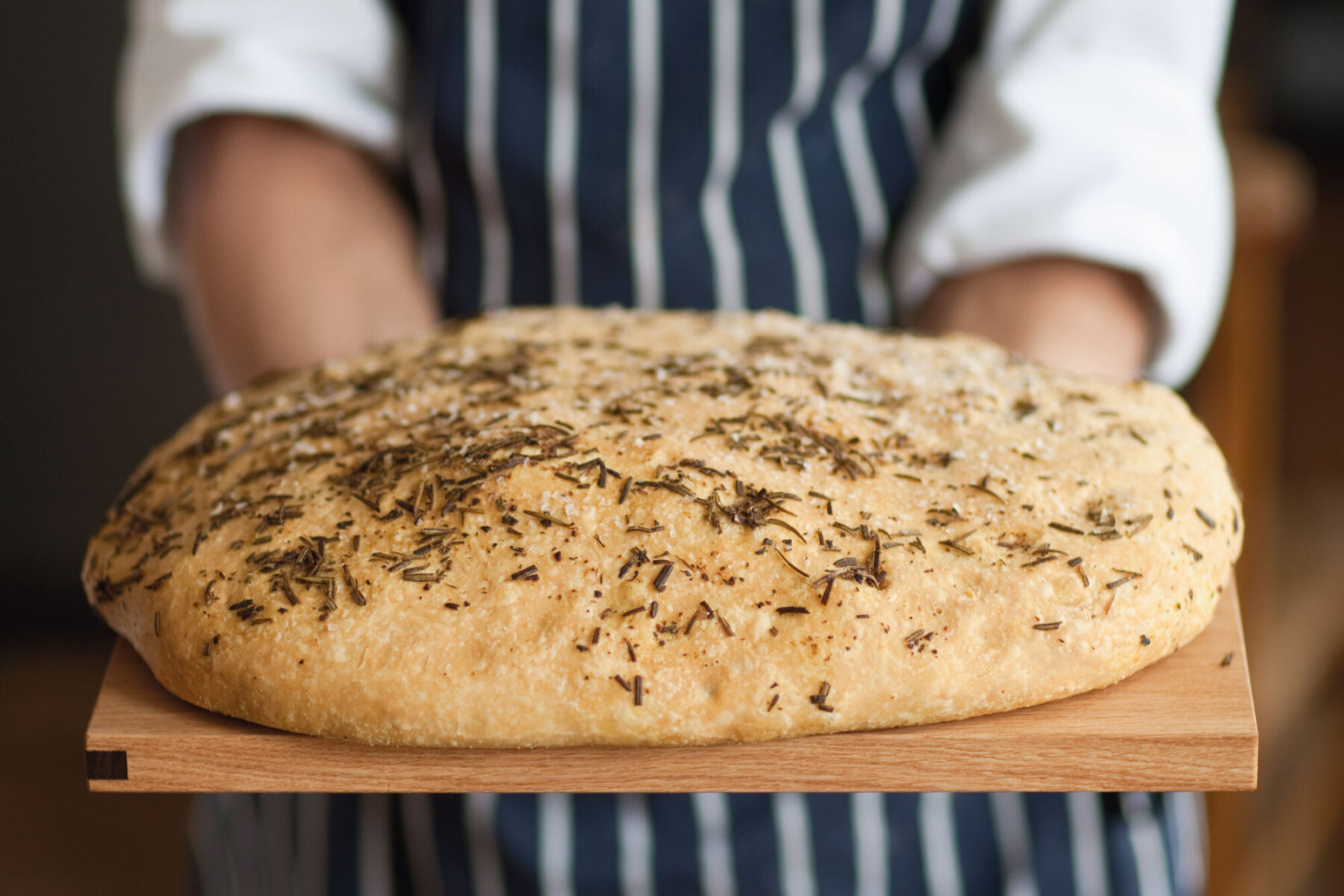 Close up of person in striped apron holding wooden board with freshly baked bread.